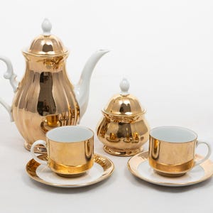 May include: A gold-colored tea set with white accents. The set includes a teapot, sugar bowl, and two teacups with saucers. The teapot and sugar bowl have matching lids. The teacups and saucers have a gold rim.