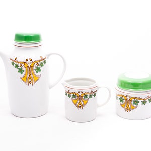 Może przedstawiać: Biały ceramiczny zestaw do herbaty z zieloną pokrywką i żółto-zielonym wzorem kwiatowym. Zestaw zawiera czajnik, dzbanek do śmietanki i cukiernicę.