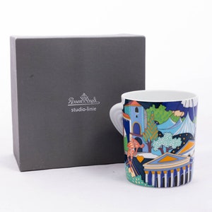 May include: A white ceramic mug with a colourful design featuring a cityscape, trees, and buildings. The mug has a handle and the text "Rosenthal studio-linie" is printed on the box.