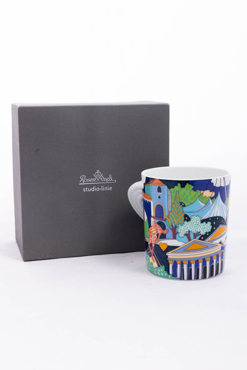 May include: A white ceramic mug with a colourful design featuring a cityscape, trees, and buildings. The mug has a handle and the text "Rosenthal studio-linie" is printed on the box.