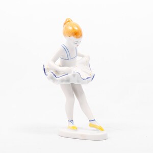 May include: A porcelain figurine of a ballerina wearing a white and blue dress with yellow shoes. The ballerina is posed with her head down and her skirt lifted.