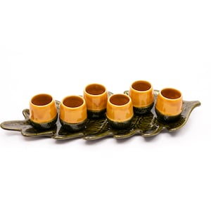 Può includere: Un set di sei piccole tazze in ceramica con smalto giallo senape e basi verde scuro, disposte su un vassoio a forma di foglia. Il vassoio è verde scuro e ha una superficie strutturata.