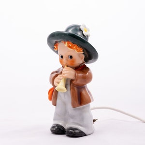 Könnte beinhalten: Eine Porzellanfigur eines Jungen, der einen grünen Hut mit einer weißen Blume trägt und eine gelbe Trompete spielt. Die Figur ist braun, weiß und orange und hat ein Kabel für ein Licht.
