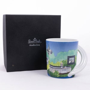 May include: A white ceramic mug featuring a blue and green cityscape design. The mug has a distinctive white handle that curves around the side. The mug is presented in a black box with the Rosenthal studio-line logo.