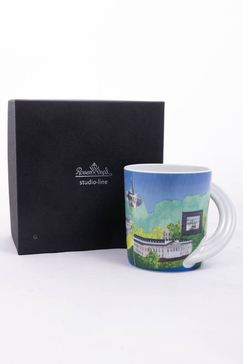 May include: A white ceramic mug featuring a blue and green cityscape design. The mug has a distinctive white handle that curves around the side. The mug is presented in a black box with the Rosenthal studio-line logo.