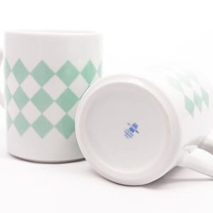 Puede incluir: Dos tazas de cerámica blanca con un patrón de diamantes verde y blanco. Las tazas tienen un asa en el lateral y un logotipo azul y blanco en la parte inferior.