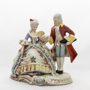 May include: A porcelain figurine depicting a couple in 18th-century attire. The woman wears a lavender and white dress with a fan, while the man is in a red coat. The base is a light beige color.