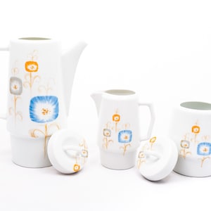 May include: A white ceramic coffee pot, creamer, and sugar bowl set with a floral design featuring blue and orange flowers. The set has a modern, minimalist style.