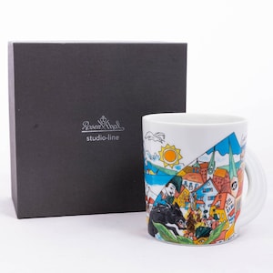 May include: A white ceramic mug with a colourful illustration of a town scene. The mug has a black handle and the words "Rosenthal studio-line" are printed on the box.