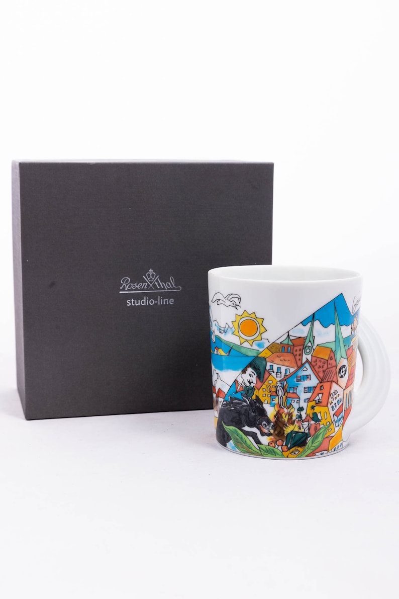 May include: A white ceramic mug with a colourful illustration of a town scene. The mug has a black handle and the words "Rosenthal studio-line" are printed on the box.