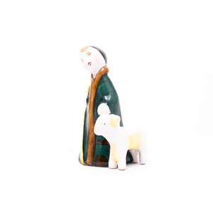 May include: A ceramic figurine of a shepherd with a green and brown robe and a white lamb. The shepherd has a painted face with brown hair and a gold trim on the robe.