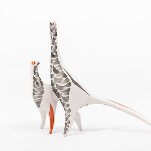May include: Two white ceramic bird figurines with black and orange accents. The birds are stylized and have long necks and tails.