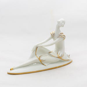 May include: A white porcelain figurine of a seated woman with gold accents. The figure is in a relaxed pose, with one leg extended. Gold trim outlines the dress and details the arms. The figurine is on a gold-edged base.