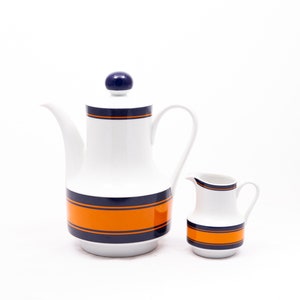 Puede incluir: Un juego de cafetera y jarra de crema de cerámica blanca con rayas azules y naranjas. La cafetera tiene una tapa con un pomo azul. La jarra de crema es más pequeña y tiene un diseño similar.