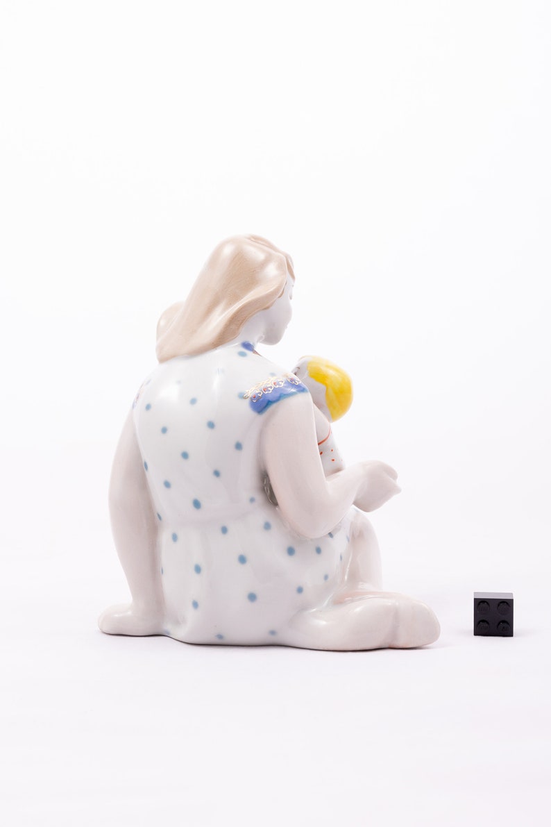 Peut inclure: Figurine en porcelaine repr&eacute;sentant une figure assise tenant un enfant. La figure assise porte une robe blanche &agrave; pois bleus. L'enfant a les cheveux jaunes. Un carr&eacute; noir est &agrave; droite.