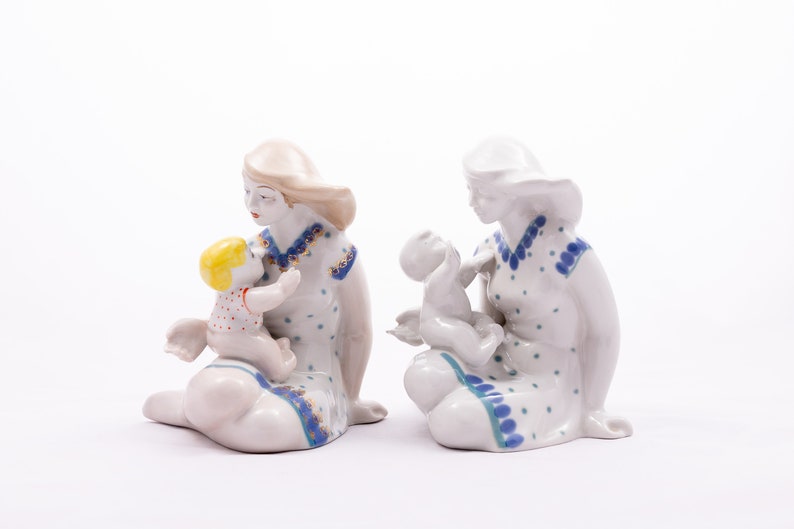Peut inclure: Deux figurines en porcelaine d'une m&egrave;re et d'un enfant. Les m&egrave;res sont assises, v&ecirc;tues de robes blanches &agrave; pois bleus et garnitures. Un enfant a les cheveux jaunes et une chemise &agrave; pois rouges. L'autre enfant est tout blanc.