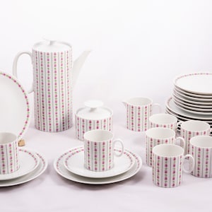 Op de afbeelding: Een wit porseleinen theeservies met een roze en groen bloemenpatroon. De set bevat een theepot, roomkan, suikerpot, kopjes, schoteltjes en dinerborden.