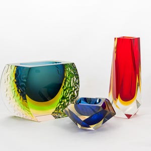 Puede incluir: Tres jarrones de cristal de colores. Un jarrón grande y rectangular con un lado texturizado verde y amarillo, y un interior azul. Un jarrón azul pequeño y facetado. Un jarrón alto y rojo con detalles amarillos.