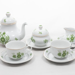 May include: A white porcelain tea set with green floral designs. The set includes a teapot, sugar bowl, creamer, and two teacups with saucers. Gold trim accents the edges of the pieces.