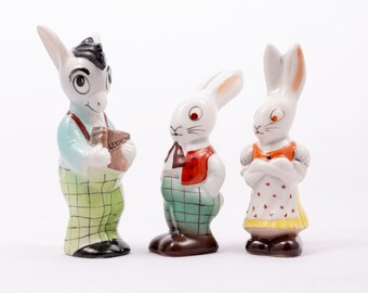 May include: Three porcelain figurines of a donkey, a rabbit, and a rabbit. The donkey is wearing a blue shirt, green plaid pants, and holding a book that says "Lexikon". The rabbits are wearing plaid pants and vests. The rabbit on the left is wearing a red vest and the rabbit on the right is wearing a yellow vest.