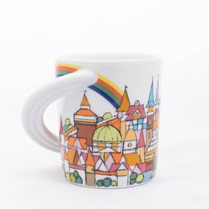 May include: White ceramic mug featuring a rainbow and a whimsical illustration of a town with colourful buildings. The mug has a unique curved handle.