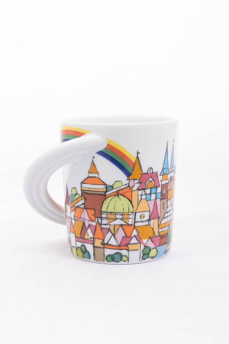 May include: White ceramic mug featuring a rainbow and a whimsical illustration of a town with colourful buildings. The mug has a unique curved handle.