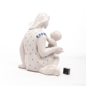 Peut inclure: Figurine en c&eacute;ramique blanche repr&eacute;sentant une m&egrave;re et son enfant. La m&egrave;re est assise, v&ecirc;tue d'une robe &agrave; pois bleus. Elle tient le b&eacute;b&eacute;. Un petit cube noir est au sol.