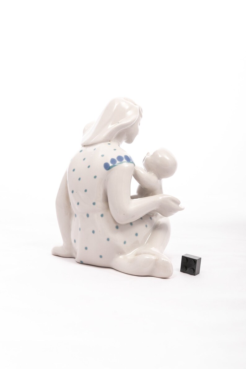 Peut inclure: Figurine en c&eacute;ramique blanche repr&eacute;sentant une m&egrave;re et son enfant. La m&egrave;re est assise, v&ecirc;tue d'une robe &agrave; pois bleus. Elle tient le b&eacute;b&eacute;. Un petit cube noir est au sol.