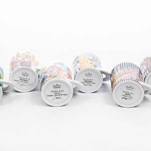 May include: Six white ceramic mugs with colourful cityscapes printed on them. Each mug has a different city name and designer printed on the bottom. The mugs are arranged in a row on a white surface.