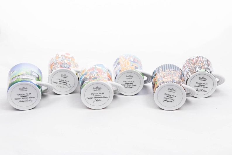May include: Six white ceramic mugs with colourful cityscapes printed on them. Each mug has a different city name and designer printed on the bottom. The mugs are arranged in a row on a white surface.