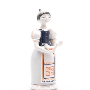 Puede incluir: Una figurita de porcelana de una mujer con un vestido azul y blanco con patrones geométricos naranja y azul. Ella sostiene un pequeño pájaro blanco en sus manos.