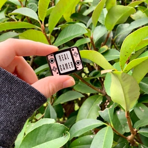 May include: A black and pink enamel pin shaped like a retro video game console with the text "REAL GAMER" on the screen.