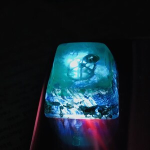 May include: A clear blue resin keycap with a silver gear mechanism inside. The keycap is illuminated from below, casting a blue glow.