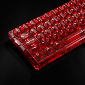 Könnte beinhalten: Eine mechanische Tastatur mit einem Gehäuse aus durchscheinendem rotem Kunststoff und passenden transparenten roten Tastenkappen. Die Tasten zeigen weiße Beschriftungen für Standard-QWERTY-Funktionen wie Esc, Tab, Caps Lock, Shift, Ctrl, Win und Alt.