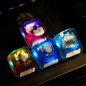 May include: Four colorful resin dice with different designs, including clouds, stars, and glitter. The dice are a vibrant purple, blue, green, and blue.