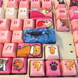 Könnte beinhalten: Eine Tastatur mit benutzerdefinierten Tastenkappen mit verschiedenen Designs, darunter ein Wal, ein Pilz, ein Hund und ein Pfotenabdruck. Die Tastenkappen sind bunt und haben eine glänzende Oberfläche.