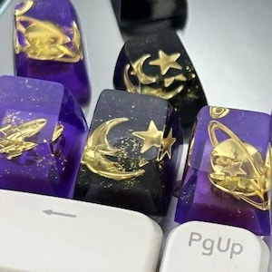 Könnte beinhalten: Nahaufnahme von individuellen Tastaturkappen in Lila und Schwarz. Die Tastenkappen zeigen goldfarbene Weltraum-Designs, darunter Raketen, Planeten, Monde und Sterne. Die Tastenkappen befinden sich auf einer weißen Tastatur.