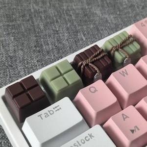 Könnte beinhalten: Nahaufnahme einer mechanischen Tastatur mit individuellen Tastenkappen in Form von Schokoriegeln, braun und grün, einige mit Bindfaden gebunden. Die Tastatur hat rosa und weiße Tasten.