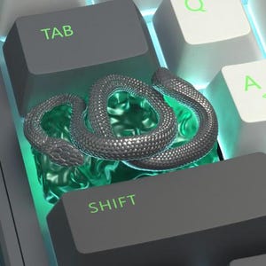 May include: A close-up of a computer keyboard with a silver snake-shaped keycap. The keycap is positioned above a glowing green light. The keyboard keys are gray and white, with green lettering.