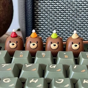 May include: Four brown bear-shaped keycaps with party hats in red, orange, green, and tan, sitting on a green keyboard. Each bear has a light brown snout and black eyes. The keyboard keys are a muted green color.