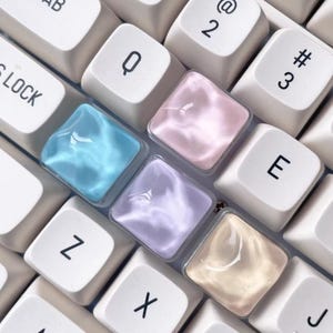 May include: Close-up of a computer keyboard with white keys and colorful, translucent keycaps. The keycaps are in shades of blue, pink, purple, and beige, creating a soft, pastel aesthetic. The keys are arranged in a standard QWERTY layout.