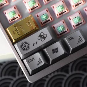 May include: A close-up of a mechanical keyboard with grey keycaps and pink switches. A gold bar is placed on the keyboard. The keycaps have various symbols, including a Windows logo, a control key, and an alt key.
