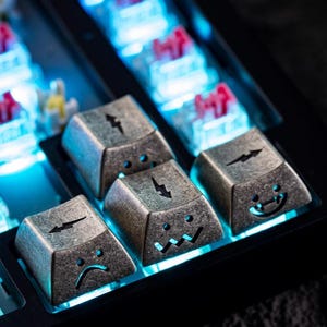 May include: Close-up of custom mechanical keyboard keycaps. The keycaps are metallic gray with etched lightning bolt and face designs. The keyboard has blue backlighting, and the keycaps have a distressed, vintage appearance.