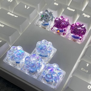 Könnte beinhalten: Ein Satz von sechs schillernden Tastenkappen mit einem Pfotenabdruck-Design. Die Tastenkappen bestehen aus klarem Kunststoff mit einem holografischen Finish. Die Tastenkappen sind in einer Reihe auf einer weißen Tastatur angeordnet.