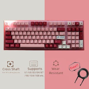 Może przedstawiać: Różowa i czerwona klawiatura mechaniczna z zestawem klawiszy, na którym widnieje napis "ダーリン!" w języku japońskim. Klawiatura ma konstrukcję z krzyżowym wałem i obsługuje układy klawiszy 61/68/83/84/87/98/104/108. Jest odporna na zużycie i jest dostarczana z wyciągarzem klawiszy.