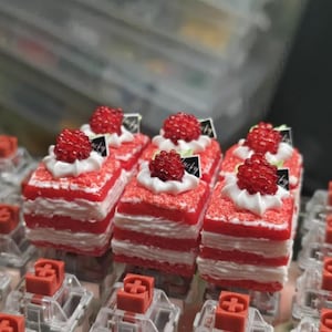 May include: Several miniature red velvet cakes with white frosting layers and a raspberry topping. Each cake has a dollop of white frosting and a small black and white label. The cakes are displayed on a surface with red and clear components.