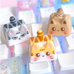 May include: Close-up of several cat-themed keycaps in various colors, including gray, brown, and orange, resting on a white keyboard. Each keycap features a unique cat design with detailed facial expressions and decorative elements. The keycaps are made of a hard, durable material.