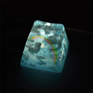 Könnte beinhalten: Eine durchsichtige blaue Tastenkappe mit einem Regenbogen- und Wolken-Design. Die Tastenkappe wird von unten beleuchtet, wodurch das Design leuchtet.