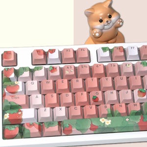 May include: A white keyboard with pink and white keycaps featuring a strawberry and leaf design. The keyboard has a white space bar with a green, white, and pink strawberry design. The keyboard is sitting on a white surface.