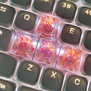 May include: Four square, translucent resin keycaps featuring embedded iridescent pink, orange, and gold glitter flakes. The keycaps are arranged in a cluster on top of a backlit computer keyboard with dark grey keys and illuminated white lettering.